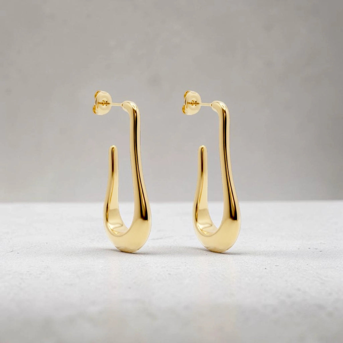 AILANA JEWELRY Kaluna earrings featuring a sculptural, elongated gold-plated stainless steel hoop design.